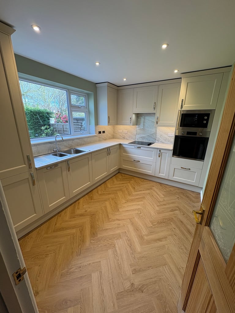 Modern white kitchen with cream cabinetry, herringbone wood flooring, stainless steel appliances, and garden window view